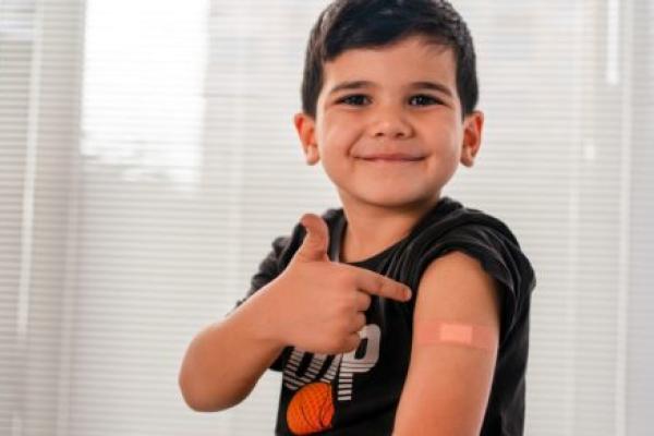 A vaccinated child points to an adhesive bandage on his arm, with his shirt sleeve rolled up.