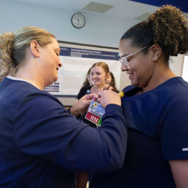 A female registered nurse attaches a pin onto the badge of a graduate nurse following the completion of her graduate nurse residency. Both are wearing navy blue scrubs. Additional graduate nurses can be seen in the background.