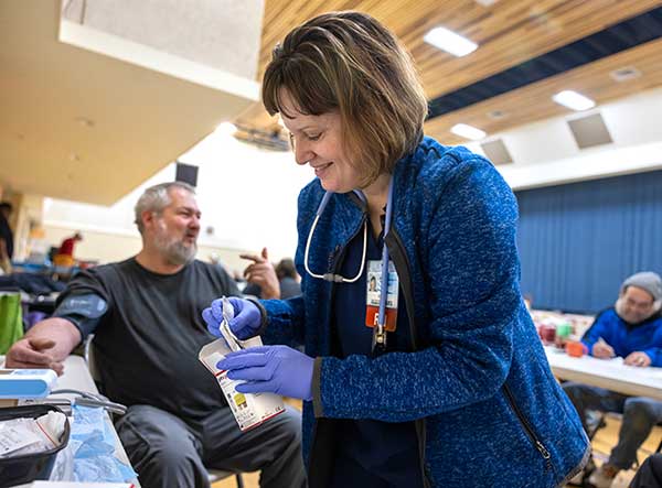 Angela Shuman smiles as she hands a man some medication. She has short hair and is wearing a jacket. The man is seated with a blood pressure cuff on his upper arm.