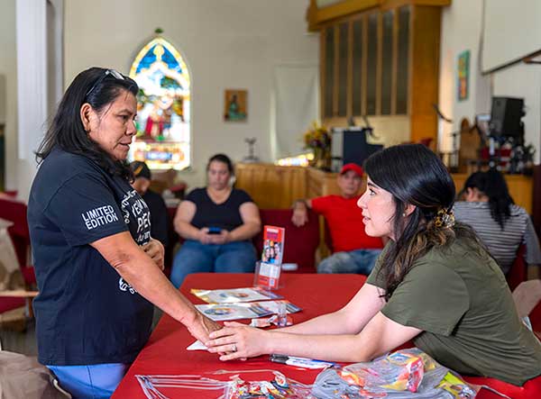 Community health worker Cristel Woodcock sits holds the hand of Maria Hernandez and looks into her eyes. Cristel has a hair clip in her long hair and is wearing a T-shirt. Maria is standing, has glasses on the top of her head and is wearing a T-shirt. Four people sit behind them.