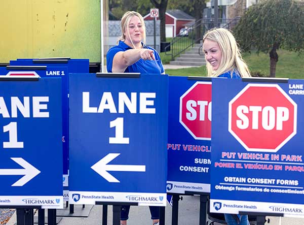 Jessica Wadsworth, left, points and Emilie Woods smiles as they stand in front of signs that read “Lane 1” and “STOP.” Behind them are steps and a tree.