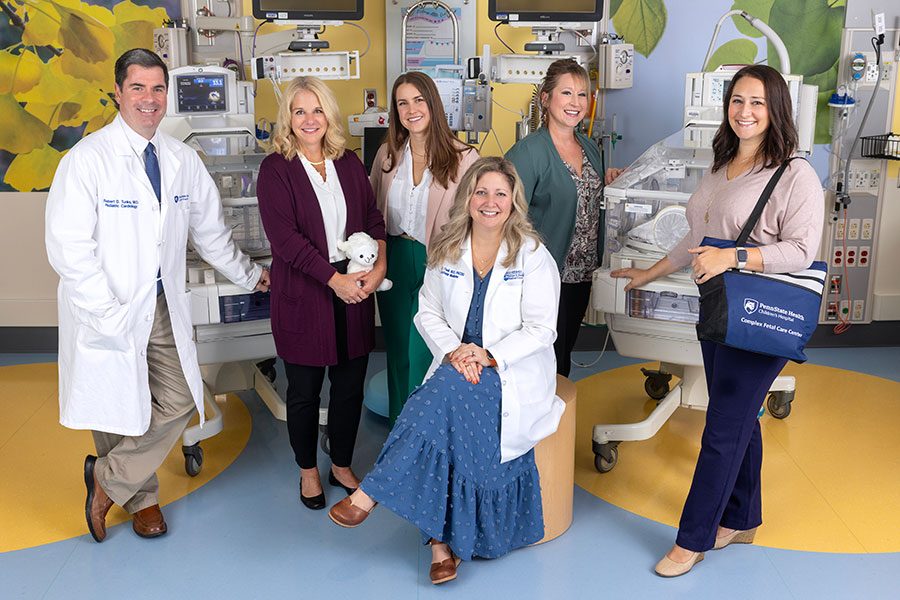 A group of medical professionals gathers in a hospital nursery. Five of them stand in the back row, while one sits in the front. The man on the left has one hand in his pocket, while he leans on an incubator. The woman standing next to him holds a stuffed animal. The woman on the far right has a bag over her shoulder. Hospital monitors and incubators are visible in the background, as well pictures of leaves on the walls.