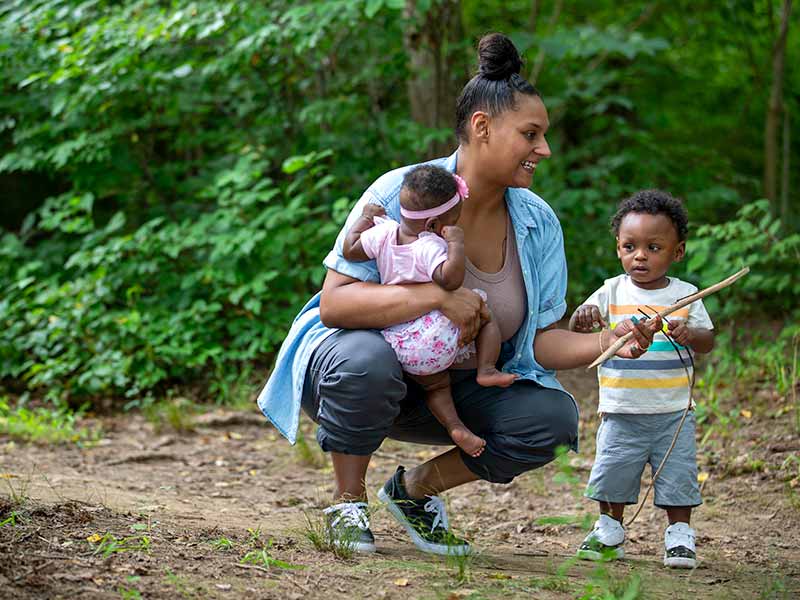 Mother squats to talk to small child while holding baby in her right arm on a wooded trail.
