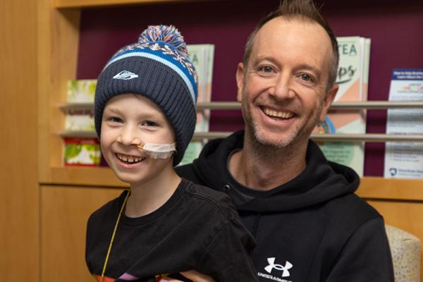 A man sits in a chair while holding a boy on his lap. The boy wears a knit cap with a pom-pom. A tube is taped to his cheek. Books sit on a shelf behind them.