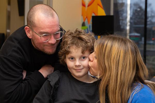 A man wearing glasses leans over the back of a chair while looking at woman kiss a little girl’s cheek. A window and monitor are visible in the background.