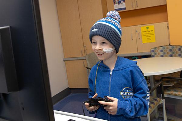 A boy wearing a knit cap with a pom-pom looks at a monitor while holding the controller for a gaming device. A tube is taped to his cheek. Behind him, a table and chairs sit in front of a wall of cabinets.