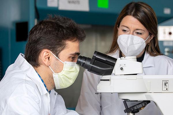 A man wearing a face mask looks into a microscope while a woman wearing a face mask watches what he is doing.