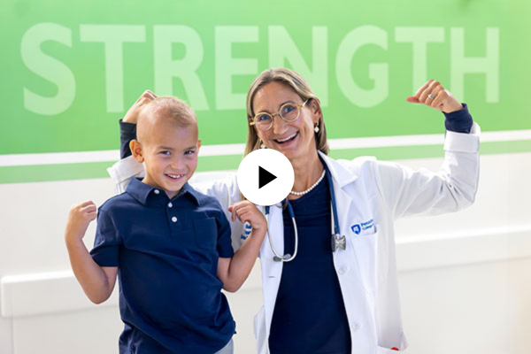 Young cancer patient and provider flexing their arms with the words strength written on a wall behind them.