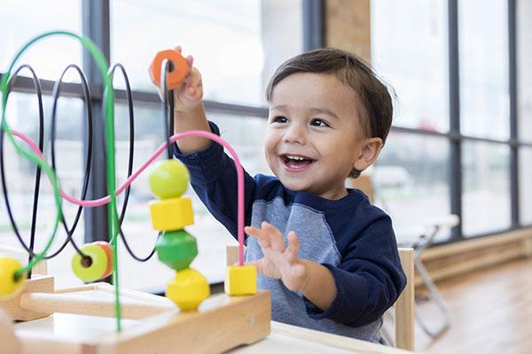 An adorable toddler boy sits at a table in a doctor's waiting room and  reaches up cheerfully to play with a toy bead maze.