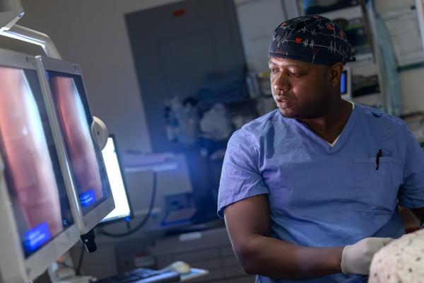 A certified registered nurse anesthetist (CRNA) in blue scrubs and a patterned cap monitors a patient's vital signs on screens during a procedure, with various medical equipment surrounding them in the operating room.]