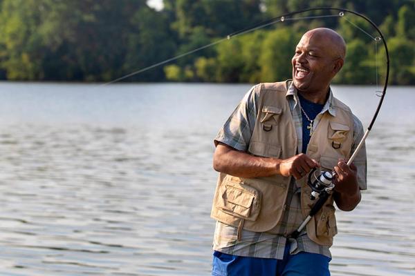 Dave, dressed in a fishing vest, stands by a lake and smiles as he reels in his fishing line. His fishing rod is bent over his head.