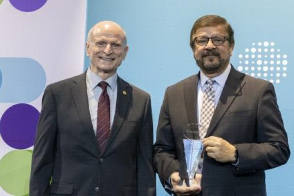 Two men stand on a stage and pose for a photo. The man on the right is holding an award.