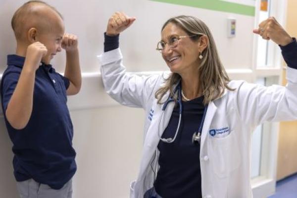 Dr. Sholler, wearing a lab coat and glasses, smiles as she raises both arms to flex her biceps. A boy next to her smiles back and mimics her.