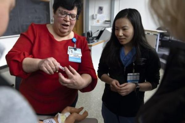 A woman in a red sweater holds a feeding tube and teaches a group of surgical residents.