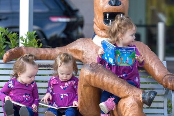 Three young girls, all dressed in identical outfits, sit on a bench with a Nittany Lion statue. One of the girls sits in the Lion’s lap, the other two are just to his side. A vehicle and a building are in the background, slightly out of focus.