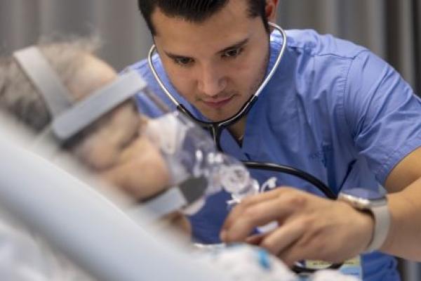 Dr. Raul Vazquez Urrutia, left, listens to the heart of a patient in the Medical Intensive Care Unit of Penn State Health Milton S. Hershey Medical Center. He has a stethoscope in his ears and is wearing scrubs. The patient is lying in a hospital bed and wearing an oxygen mask.