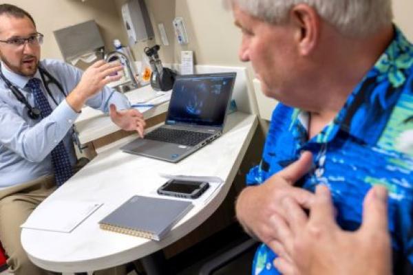 Tyler Thomas, left, gestures as he talks to patient John Jones, right. Thomas is wearing a button-down shirt, tie, glasses and has a stethoscope around his neck. Jones is wearing a Hawaiian shirt and has his hands over his heart.