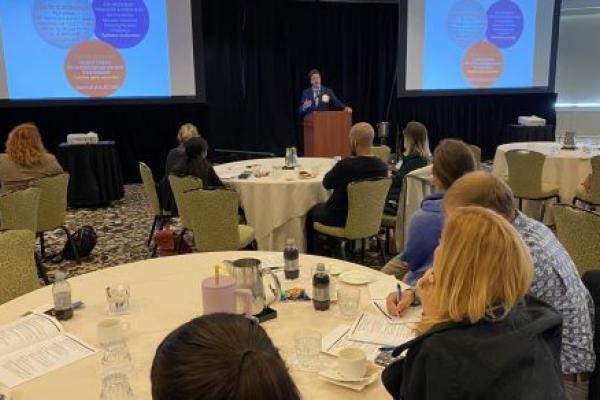 Dr. Fabio Cominelli stands at a podium, flanked on both sides by projector screens, in a room full of people seated at tables.