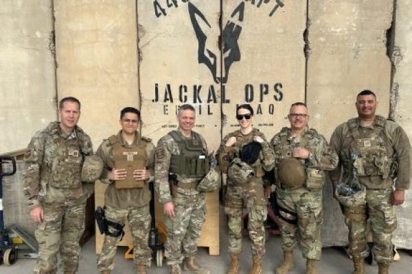 Six U.S. military personnel in camouflage stand together outside, wearing tactical gear, in front of a wall with a “Jackal Ops” logo and “Iraq.”