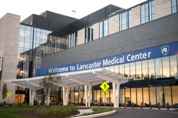 Front entrance of Penn State Health Lancaster Medical Center. The building features a sleek design with extensive glass windows, beige and dark gray brickwork, and a covered entrance with white, curved support beams. A pedestrian crossing sign is visible near the driveway, surrounded by green landscaping and trees.