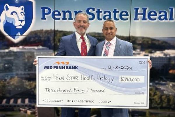 Two men in suits pose with a check made out to “Penn State Health Urology” in the amount of $340,000. A large Penn State Health logo is in the background.