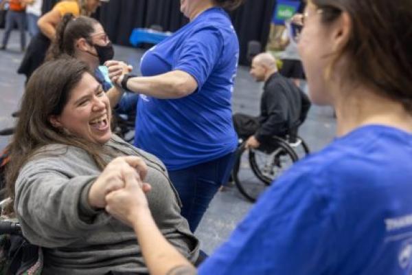 Two women, one standing the other in a wheelchair, hold hands and smile while dancing. Other dancers are in the background.