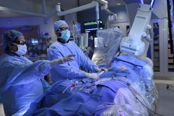 Two physicians perform a procedure on a draped patient in a surgical setting. Various pieces of medical equipment are located throughout the room.