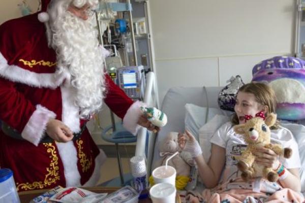 A young girl in a hospital bed, in a room filled with medical equipment and other supplies, smiles as she reached out to accept a gift of a small stuffed animal from Santa Claus.