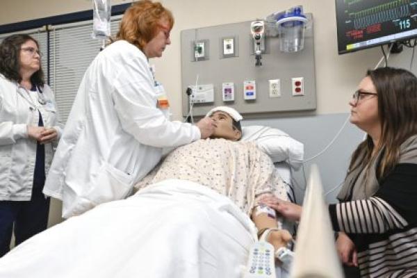 Two nurses tend to a mannequin posing as a patient while a woman sits at the bedside, looking on.