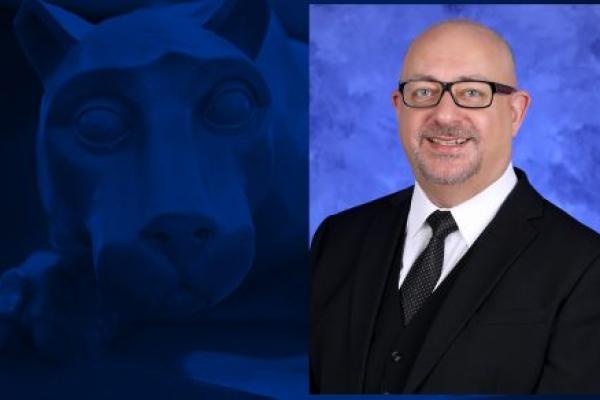 A headshot of a man smiling is overlaid on a blue background that has a statue of a lion mascot