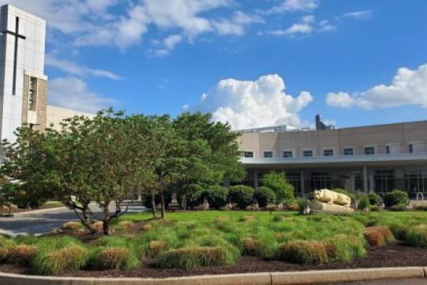 Front entrance of Penn State Health St. Joseph Medical Center with a landscaped circular driveway, a grassy area featuring small bushes, trees, and a statue of the nittany lion. The building has a tall structure with a cross, large glass windows, and a blue sky with scattered white clouds in the background.