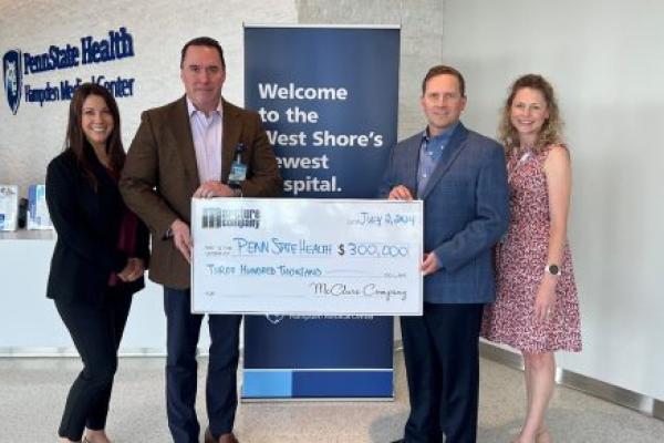 Two men and two women pose in front of a vertical banner that says, “Welcome to the West Shore’s newest hospital.” The gentlemen are in the middle holding a large presentation check for $300,000 written to Penn State Health from McClure Company.