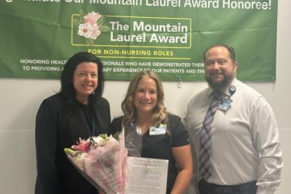 Two ladies and a gentleman stand side-by-side, smiling. The lady in the middle holds a certificate and flowers. On the wall above and behind them is a banner that says, “Congratulate Our Mountain Laurel Award Honoree! The Mountain Laurel Award for non-nursing roles.”