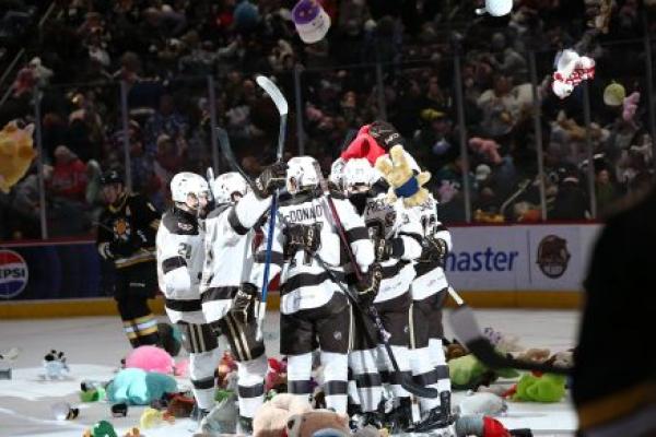 A group of professional hockey players gather facing each other on a hockey rink. Stuffed animals are on the ice around them and flying through the air from the spectators who are throwing them.