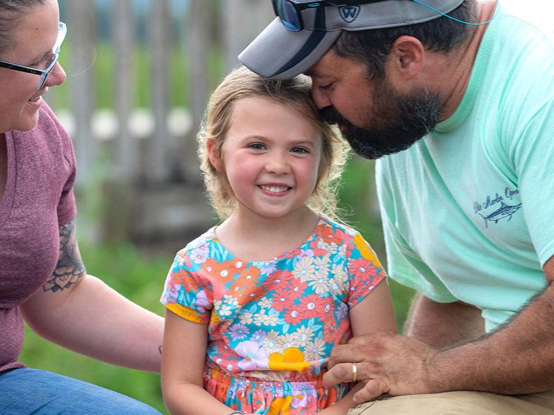 A family sits on a park bench. The father is leaning down kissing the daughter on the head and the mother is smiling at the daughter.