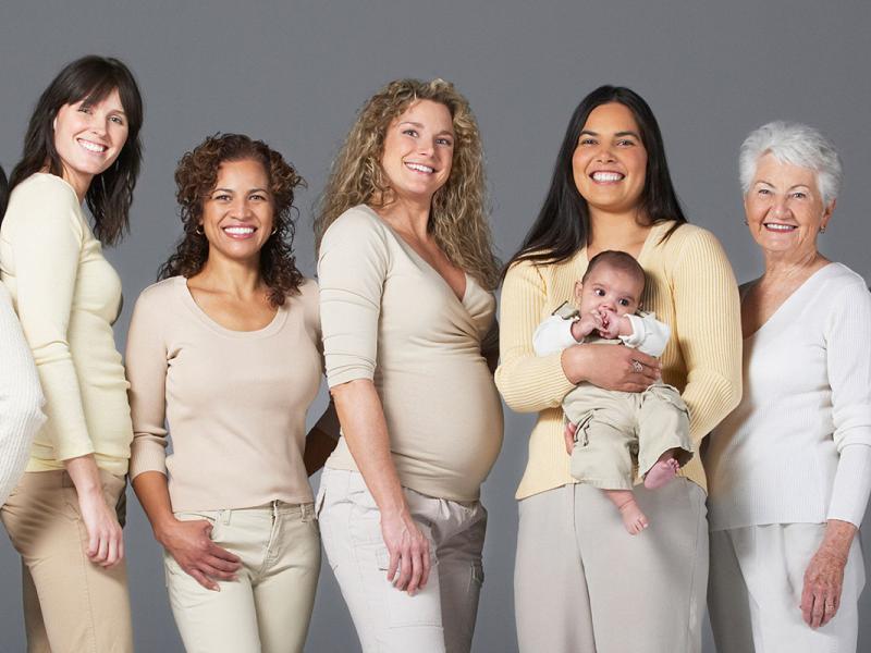 A diverse group of smiling women stand side by side and some back to back. One woman holds a baby.