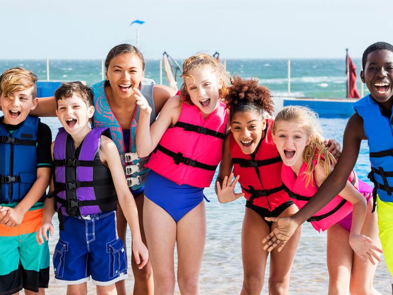 A group of children in life vests stands with their teacher in front of the ocean. Their mouths are open in big smiles and some of them are waving their arms.