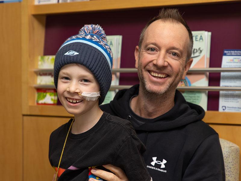 A man sits in a chair while holding a boy on his lap. The boy wears a knit cap with a pom-pom. A tube is taped to his cheek. Books sit on a shelf behind them.