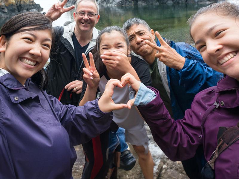 A group of male and female hikers smile at the camera while making the “peace” sign with their hands. The two female hikers in the front bring their hands together to form a “heart” sign. The female hiker in the center of the photo holds her hand over her mouth. Behind the hikers, rocks and trees rise above the water.