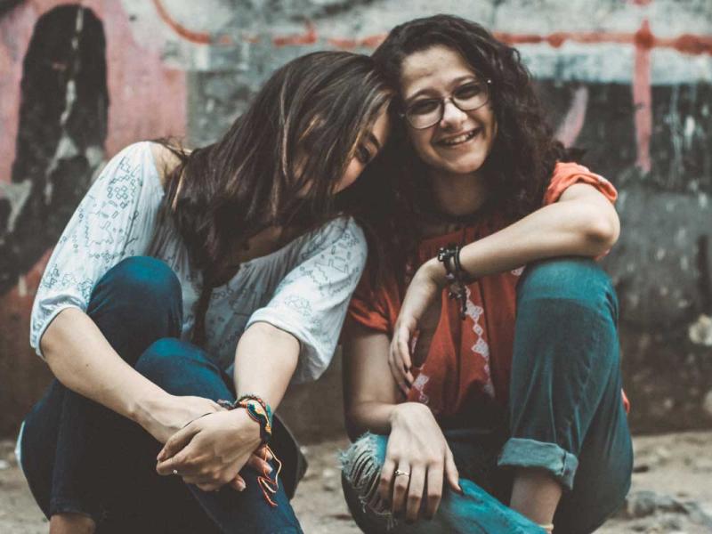 Two Girls Sitting on Pavement Near Painted Wall