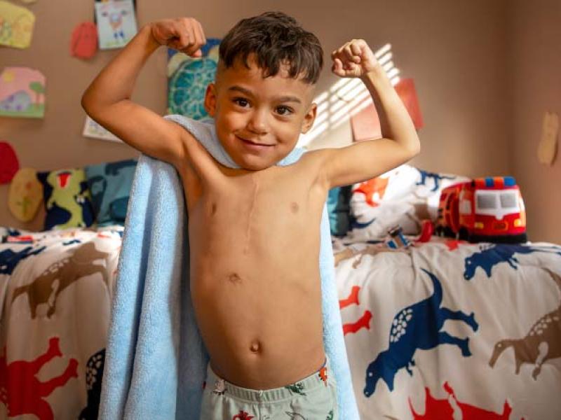 JuJu Rivera, a five-year-old boy from Lebanon who received three heart surgeries at Penn State Children’s Hospital, makes muscles with both arms as he stands in his bedroom. He has a vertical scar on his chest and is wearing a towel on his back like a superhero cape and pajama pants. Behind him, his bed is covered with a bedspread decorated with dinosaurs.